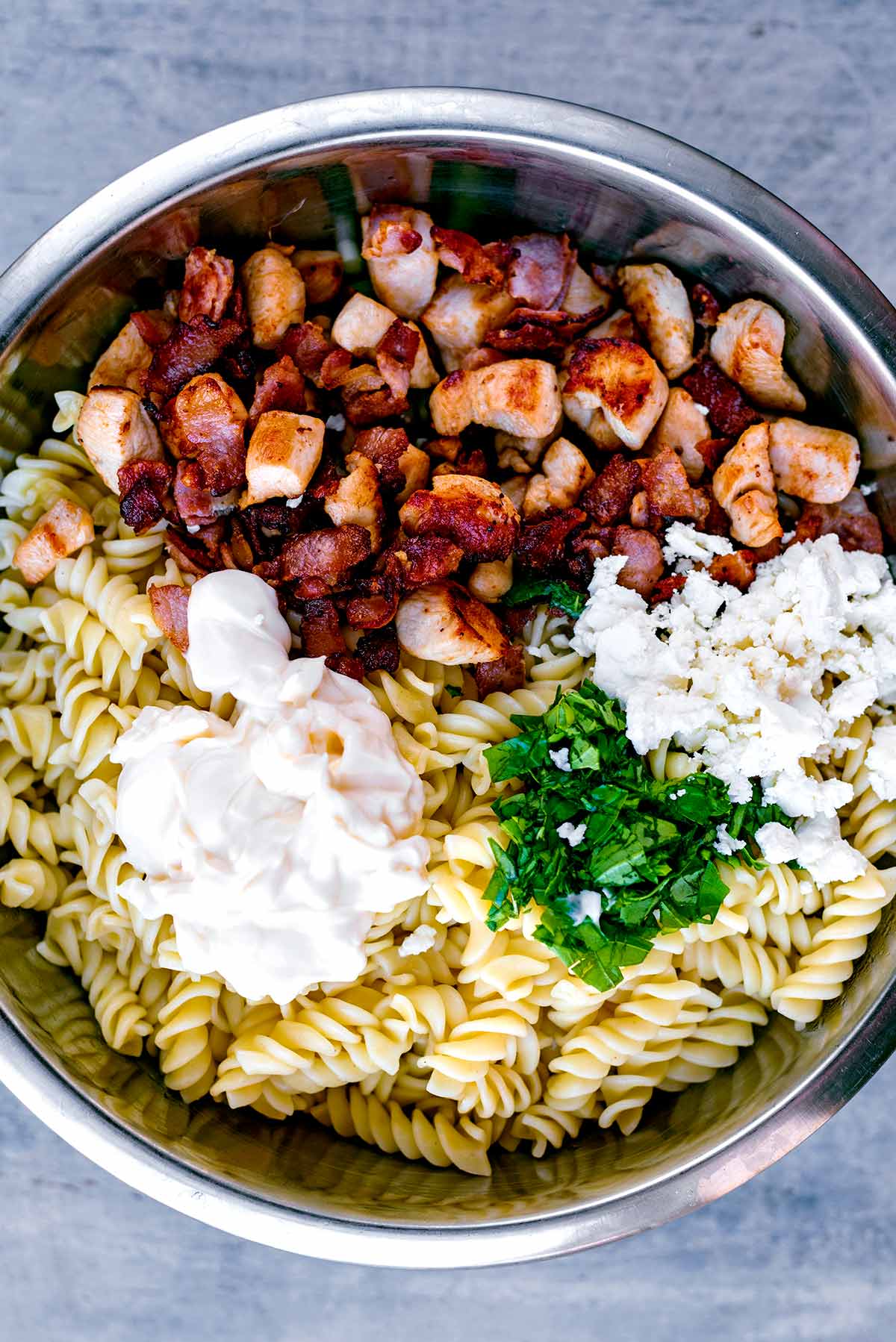 Chicken, bacon, pasta, cheese, salad vegetables, herbs and mayonnaise in a bowl.