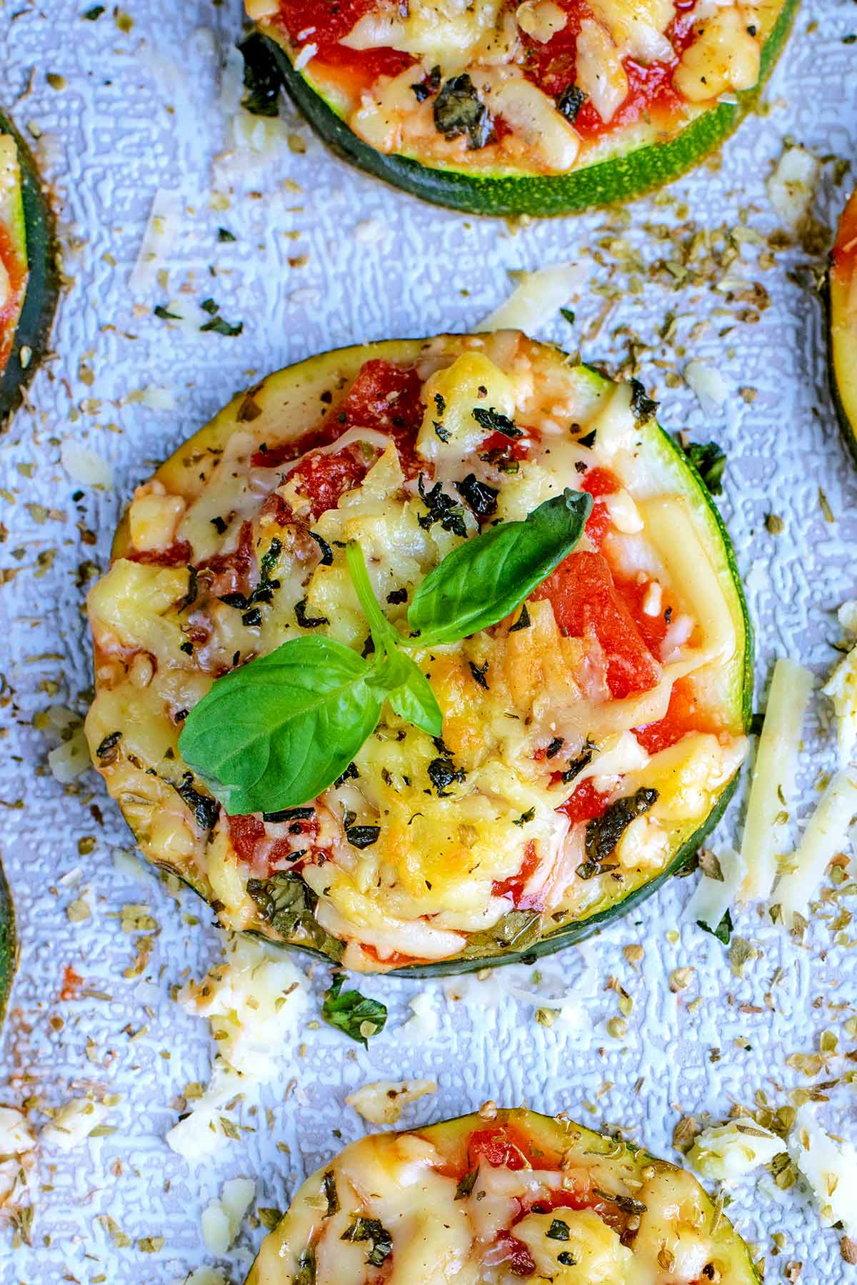 A large slice of courgette topped with chopped tomato, melted cheese and basil leaves.