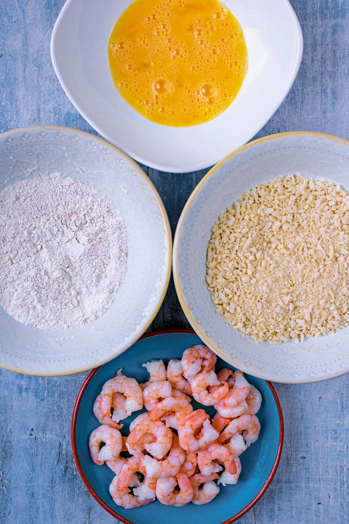 A plate of prawns, and bowls of flour, breadcrumbs and whisked egg.