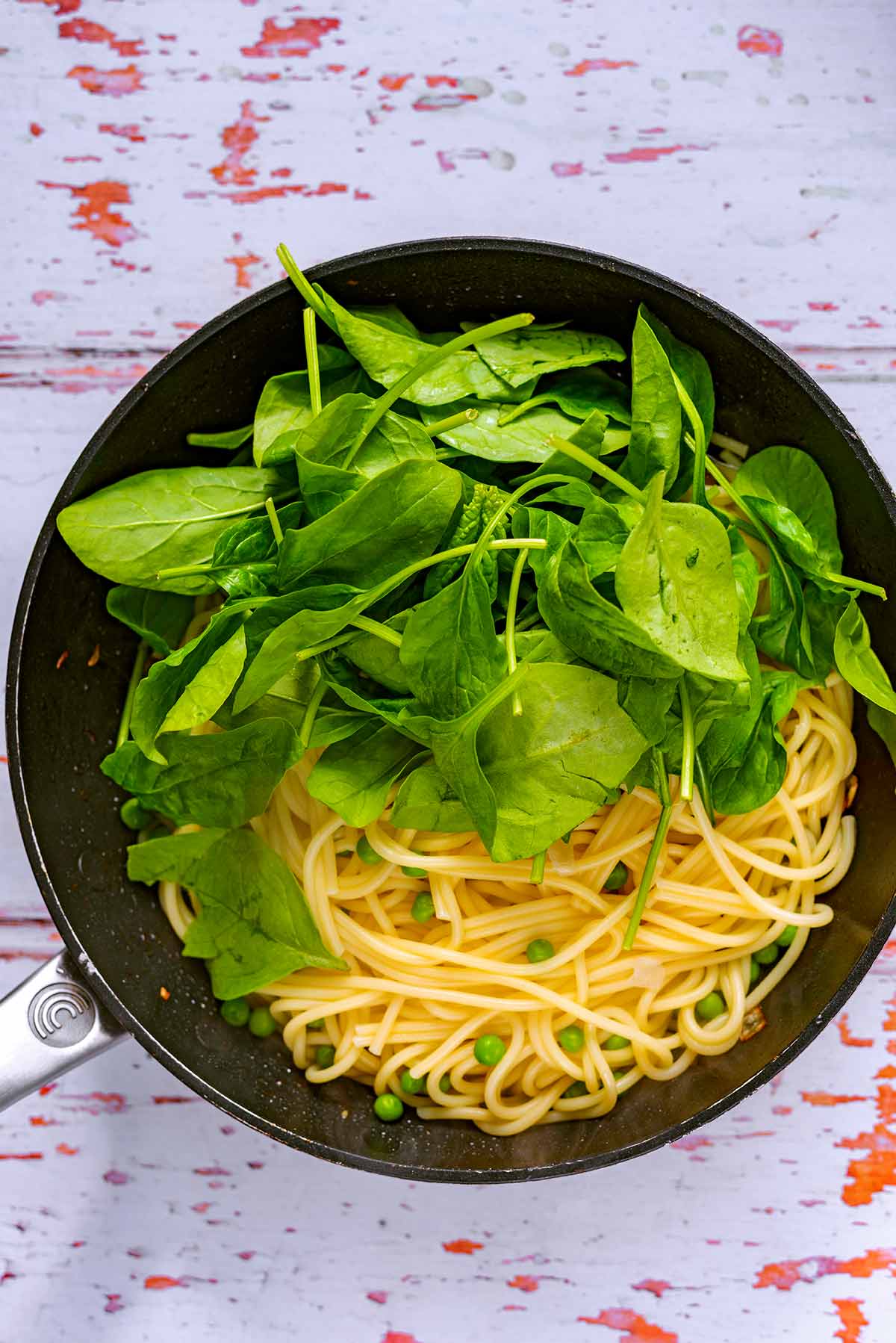 A frying pan containing spaghetti, bacon, spinach and peas.