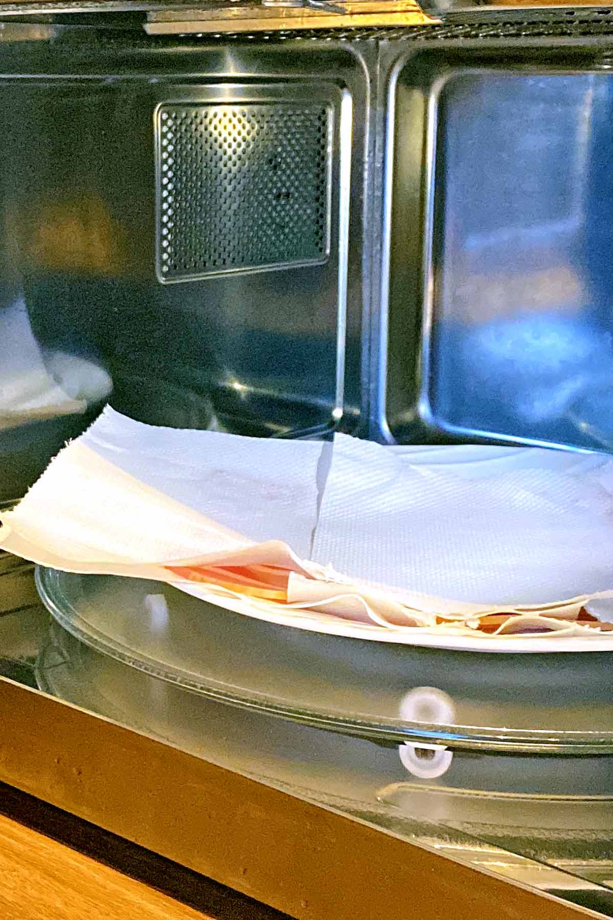 An open microwave with a plate of raw bacon covered in kitchen paper.
