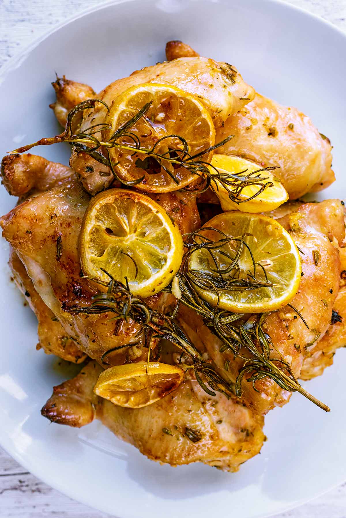 Four baked chicken thighs on a plate with lemon slices and sprigs of rosemary.