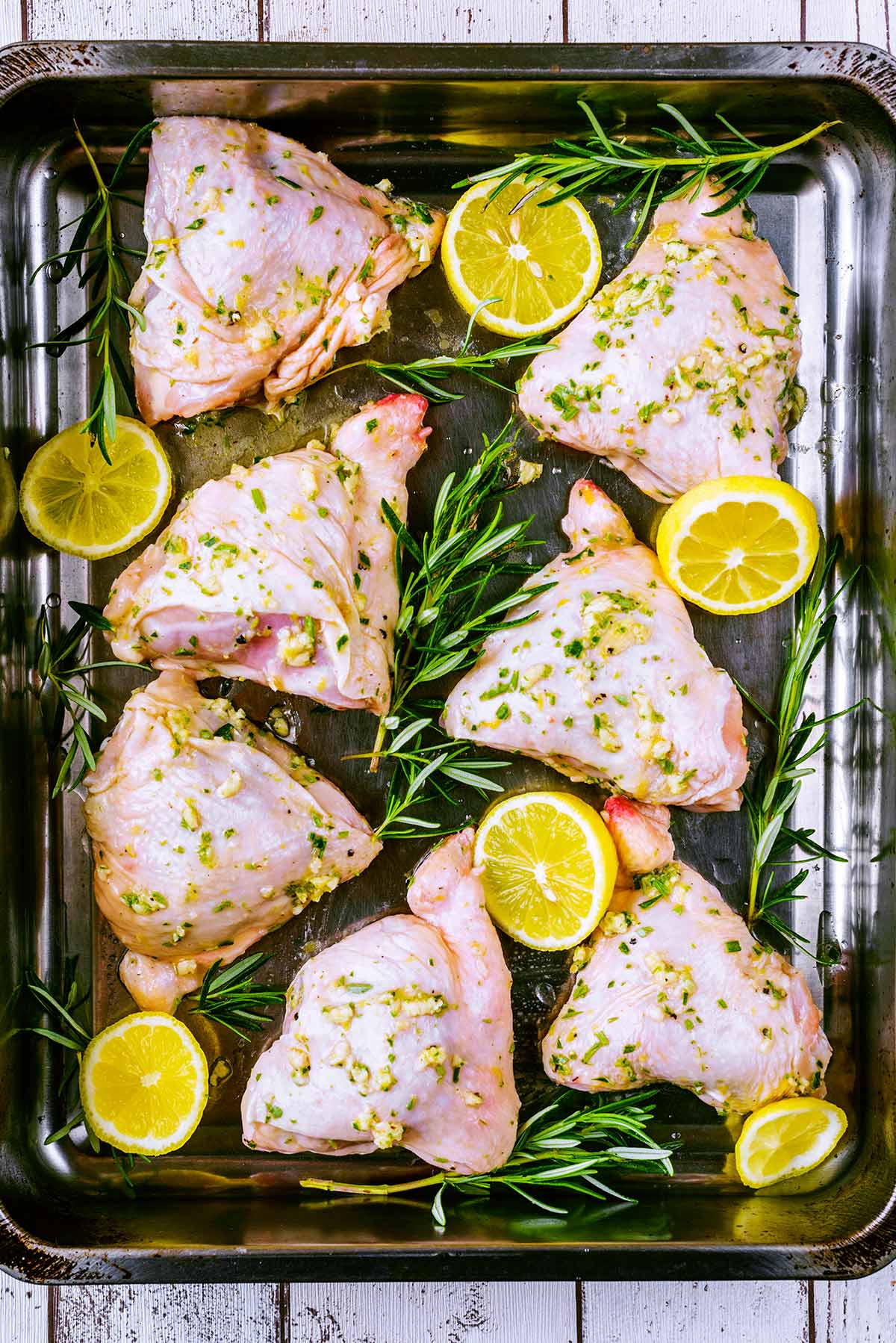 A roasting tin containing chicken thighs lemon slices and sprigs of rosemary.