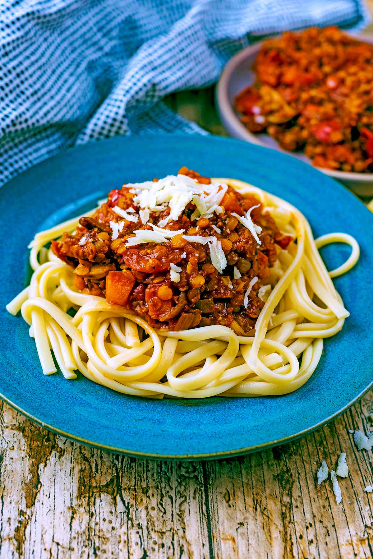 A plate of pasta and lentil bolognese in front of a spotted towel and more bolognese.