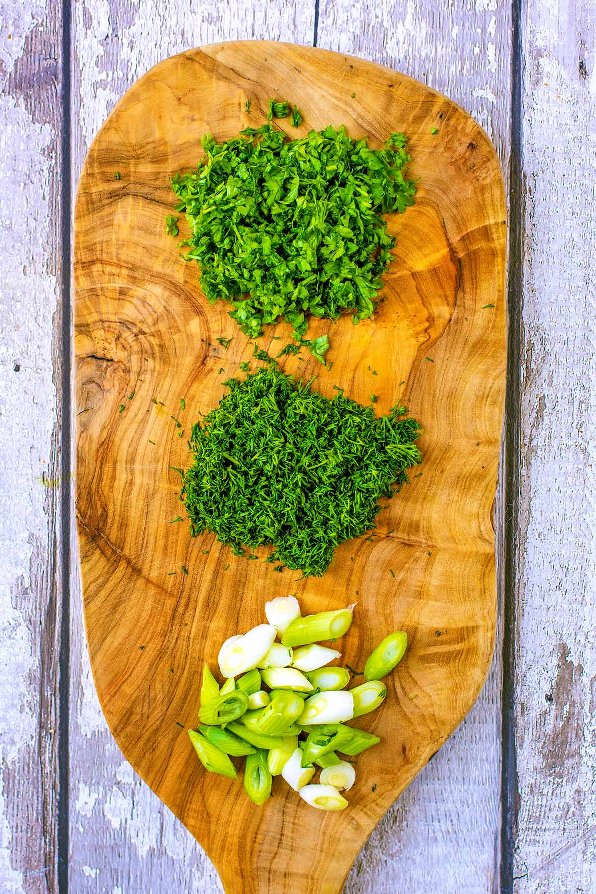 A wooden chopping board with chopped herbs and spring onions on it.