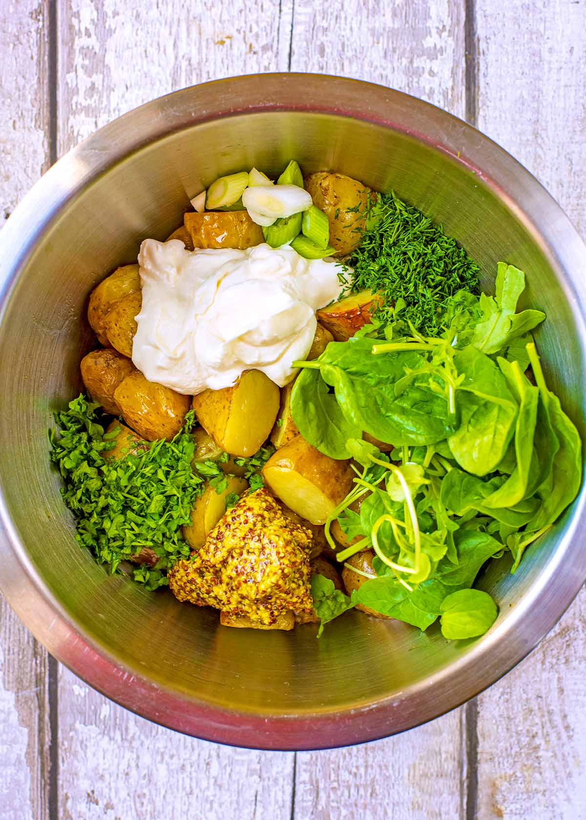 A large mixing bowl containing potatoes, salad leaves, mustard, herbs and creme fraiche.
