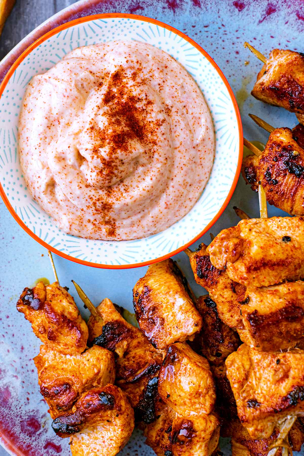 A bowl of creamy dip with paprika sprinkled over it. Chicken skewers sit next to it.