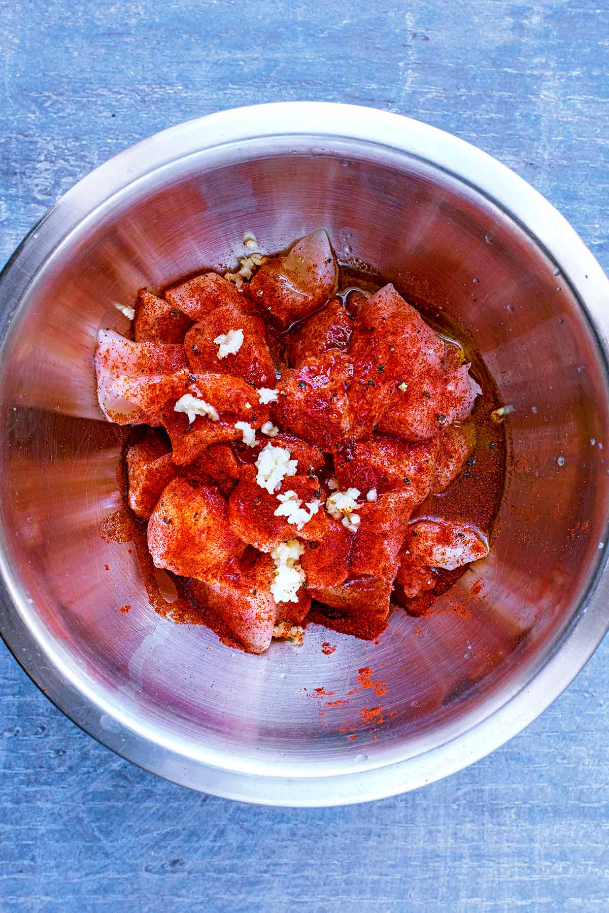 Chunks of raw chicken breast and seasoning in a metal mixing bowl.