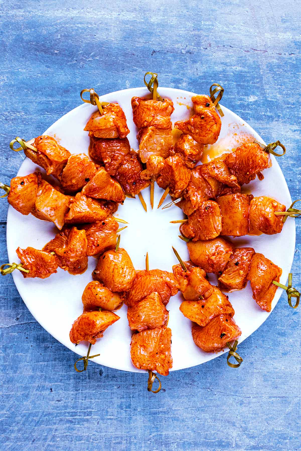 12 uncooked chicken skewers arranged in a circle on a white plate.