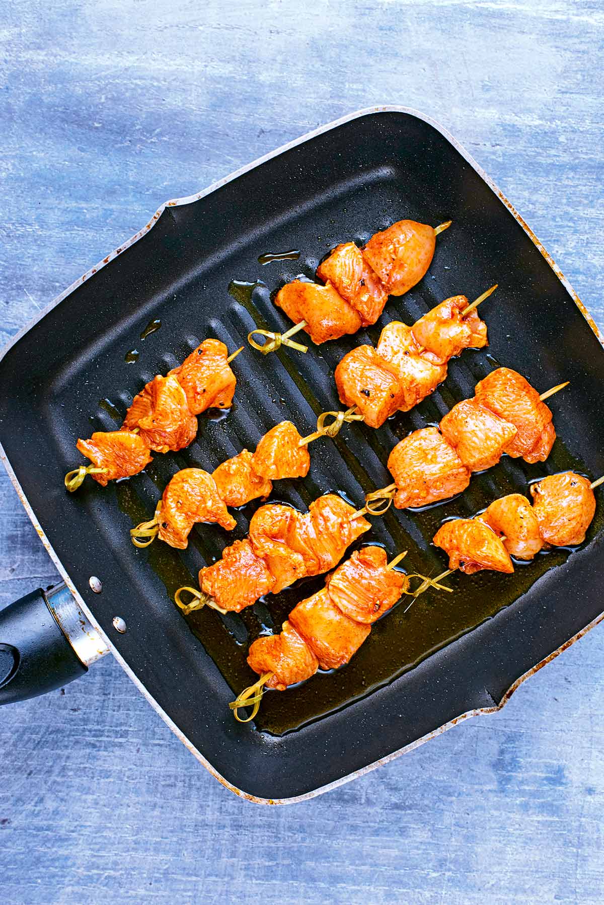 A square griddle pan with eight uncooked chicken skewers in it.