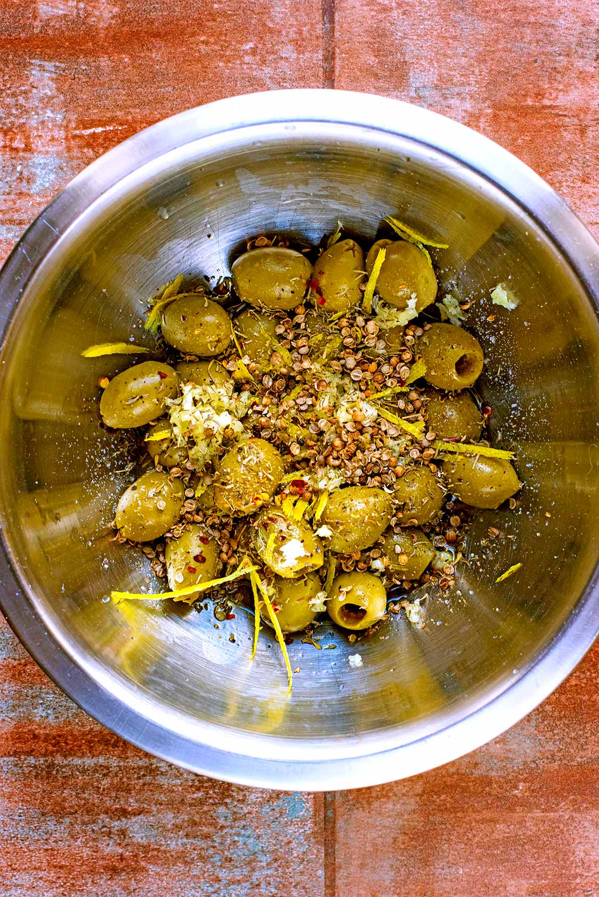 A metal bowl containing green olives marinated in oil, herbs and lemon zest.