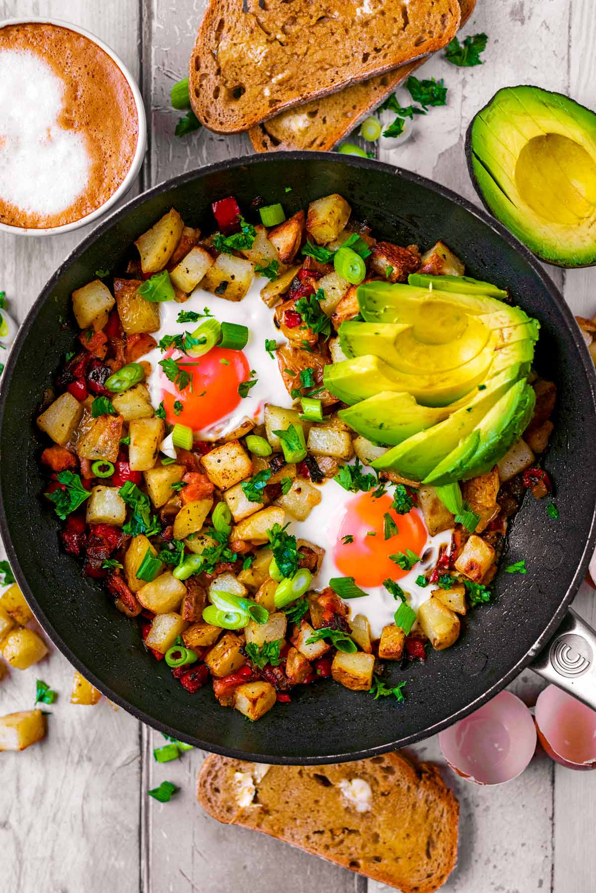A pan of breakfast hash next to toast, coffee and an avocado.