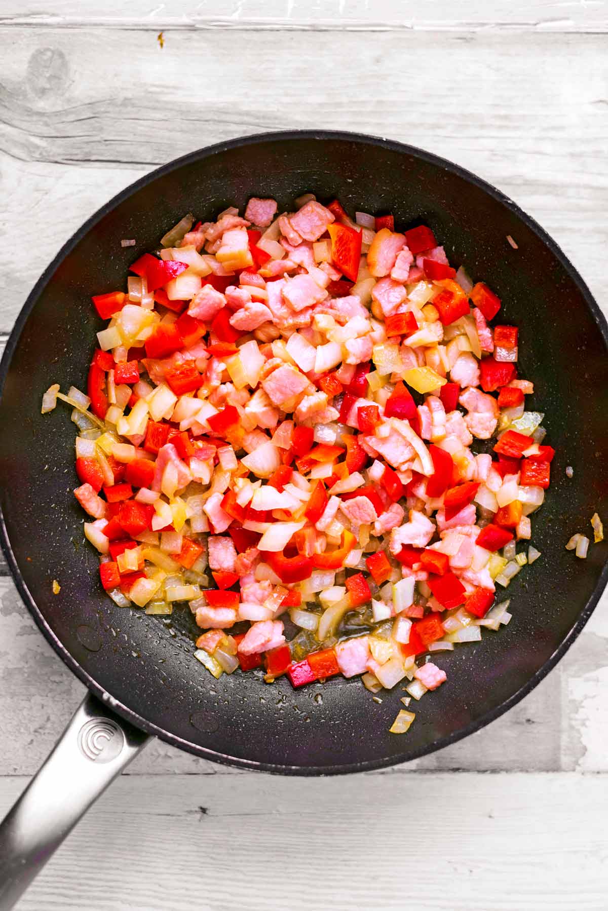 A frying pan with onion, red pepper and bacon cooking in it.