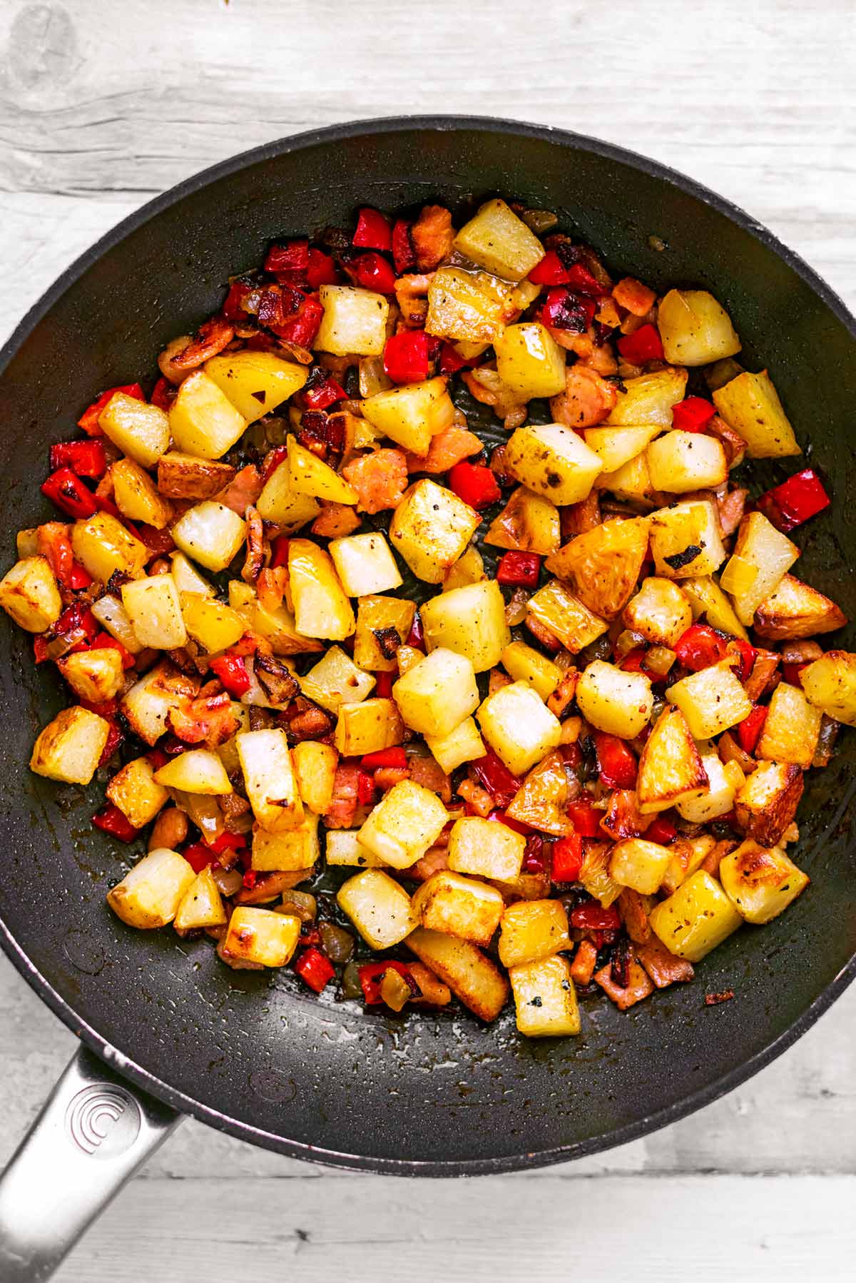 A frying pan with cubed potatoes, vegetables, bacon and spices cooking in it.