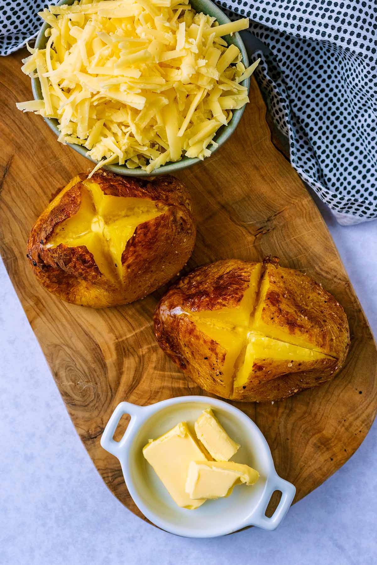 Two jacket potatoes, some grated cheese and a dish of butter on a serving board.