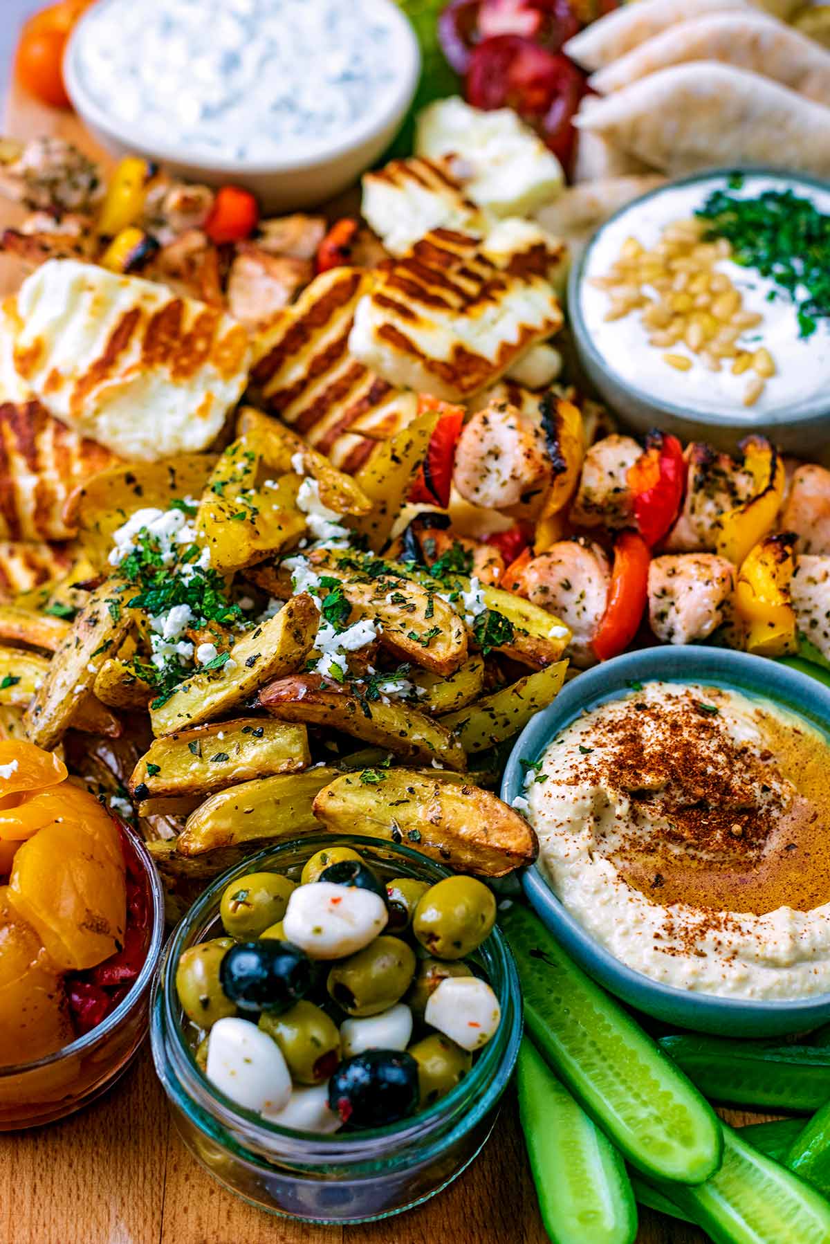 A wooden board covered in dips, vegetables, grilled halloumi slices, olives and chicken skewers.