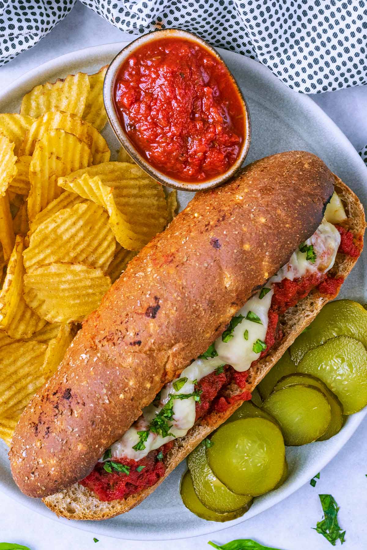 A meatball sub on a plate with crisps, pickles and a small bowl of marinara.
