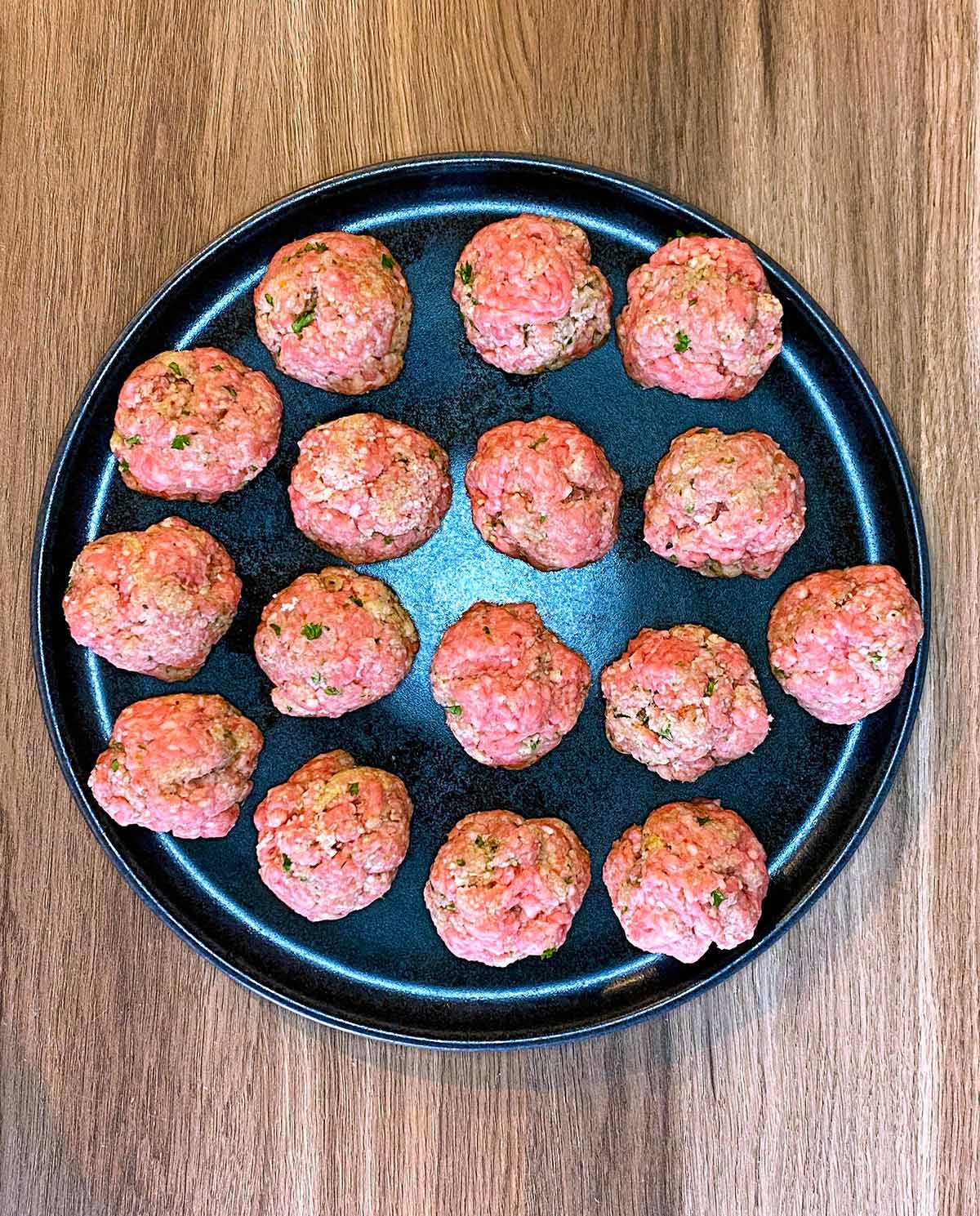 Sixteen uncooked meatballs on a black plate.