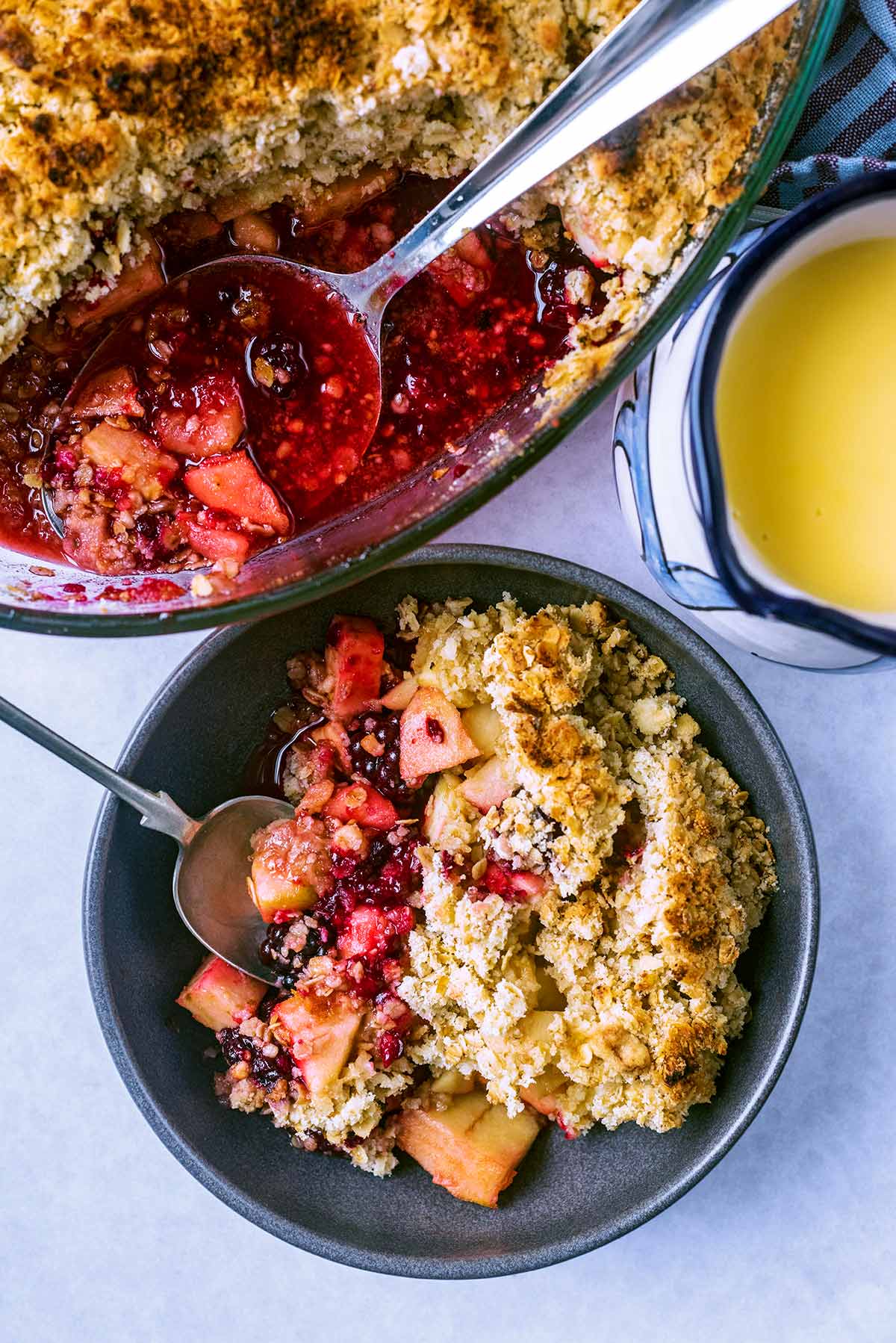 A bowl of fruit crumble next to a dish of more crumble and a jug of custard.