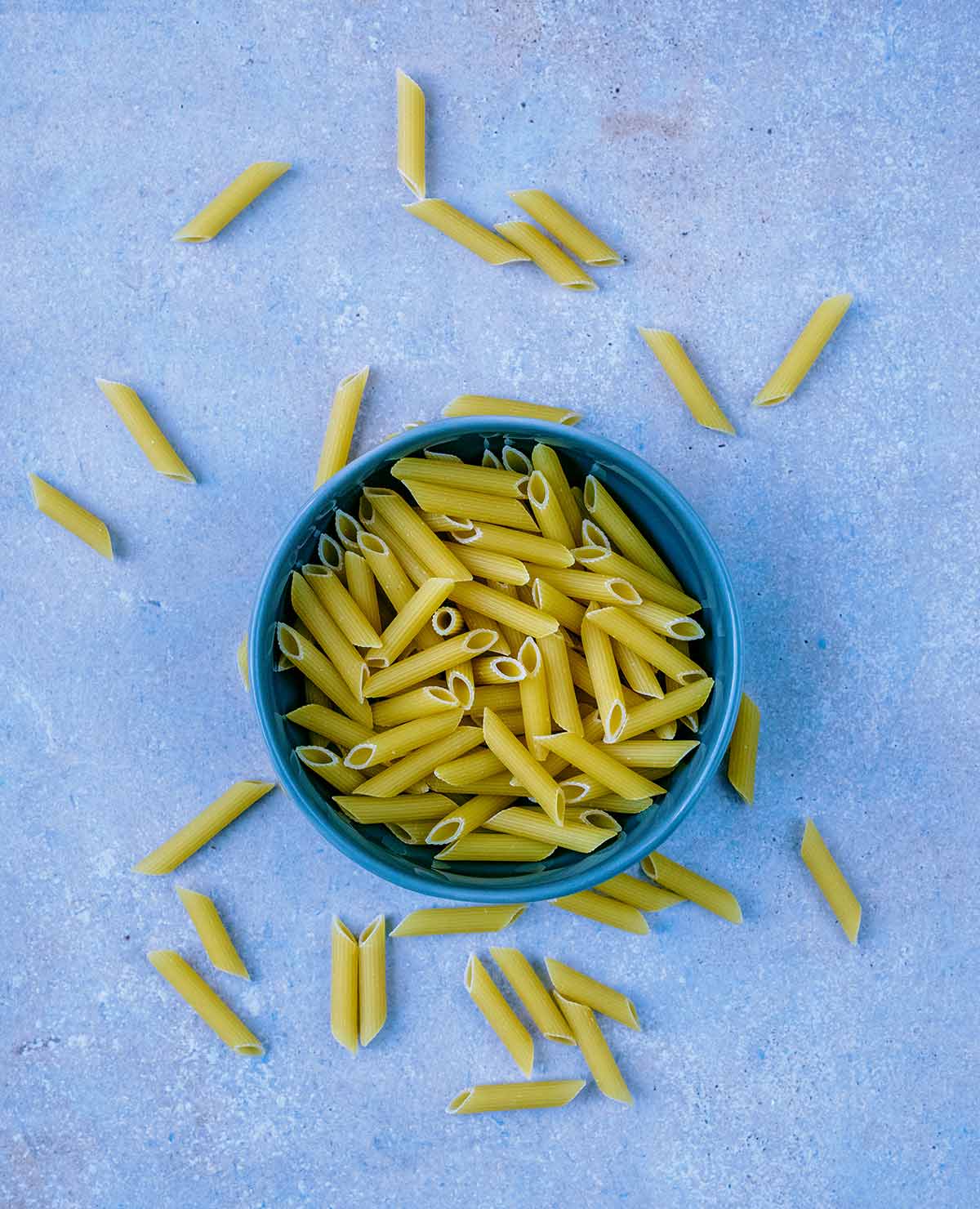 A bowl of uncooked penne pasta with more pasta scattered around it.