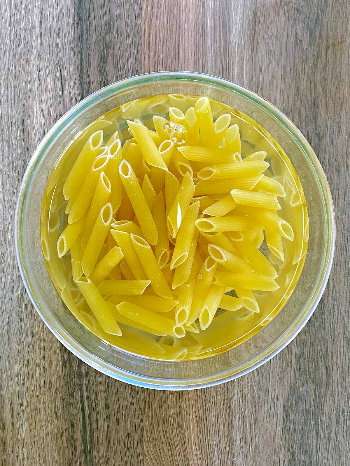 Uncooked pasta in a glass bowl covered in water.