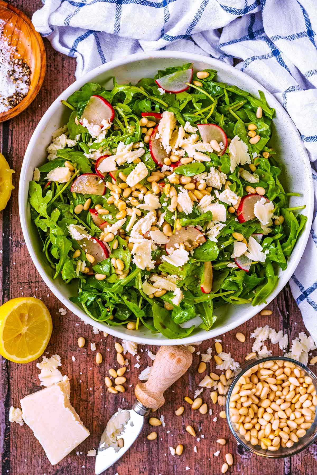 A large bowl of salad next to a towel, some pine nuts and a block of Parmesan cheese.