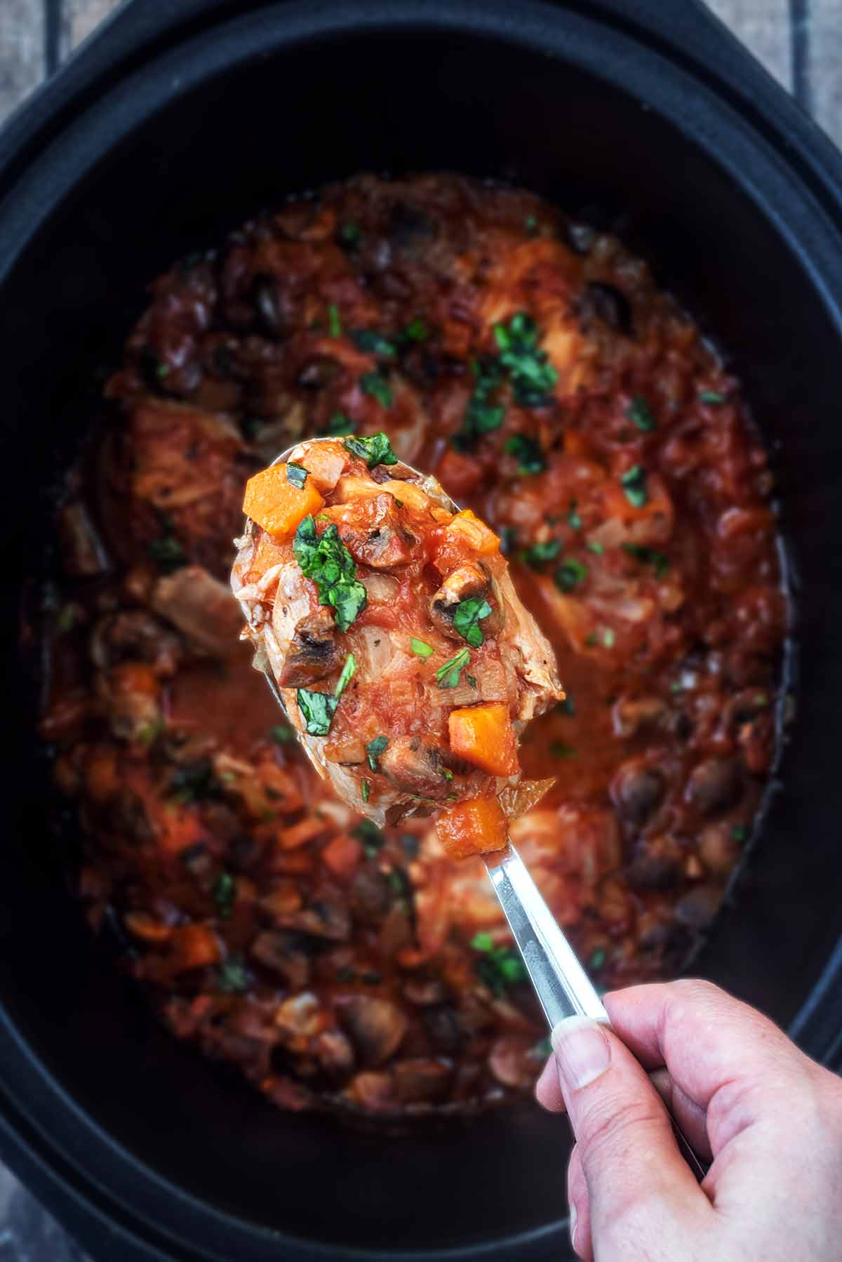 A spoon lifting chicken chasseur from a slow cooker bowl.