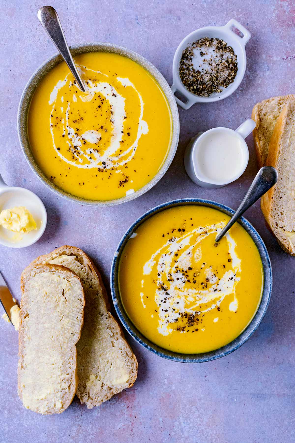 Two bowls of vegetable soup next to some buttered bread and a small jug of cream.