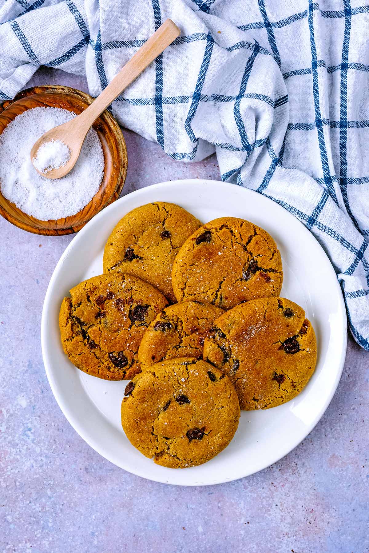 Six chocolate chip cookies on a plate next to a towel and dish of salt.
