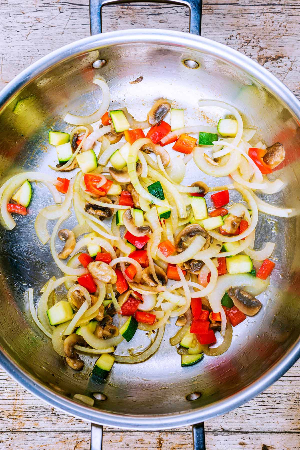 A large silver pan with onions, mushrooms, peppers and zucchini cooking in it.