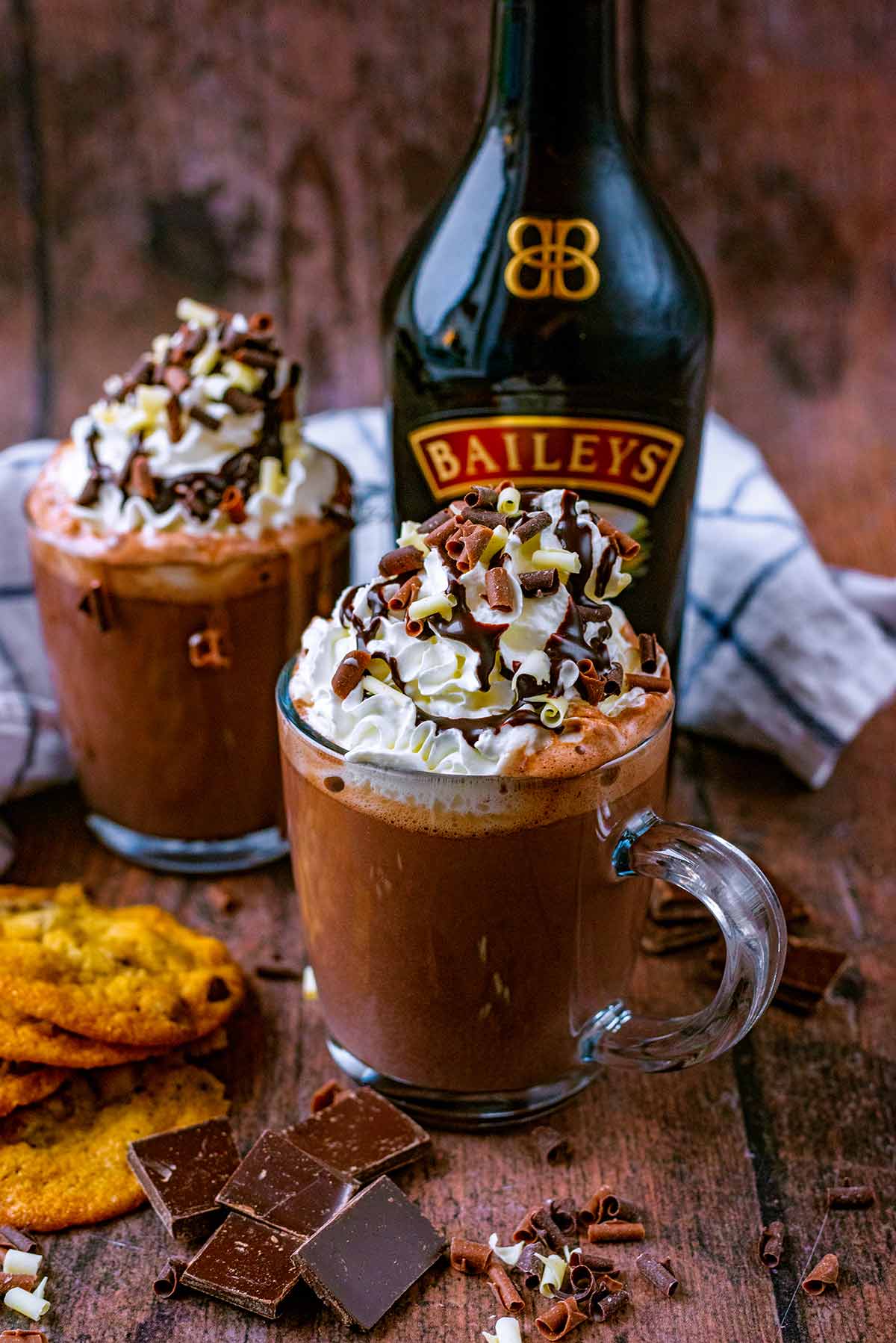 Two glasses of cream topped hot chocolate in front of a bottle of Baileys.