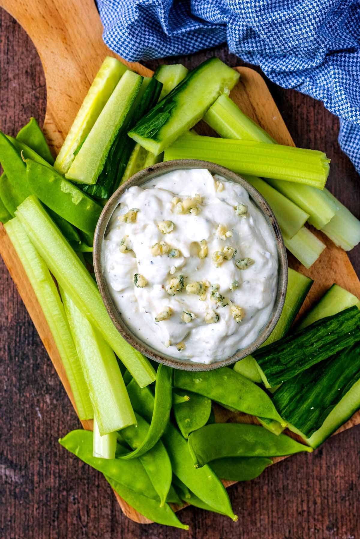 A bowl of creamy dip on a board with cucumber, celery and mange tout.