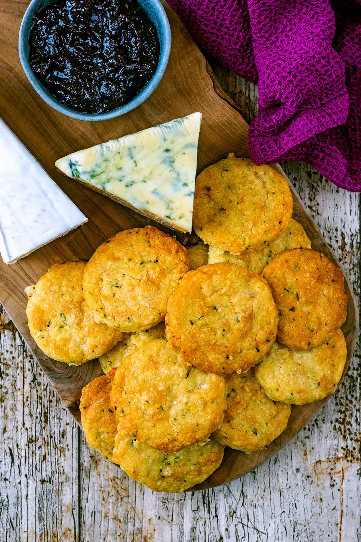 A pile of cheese biscuits on a wooden board with cheese and chutney.