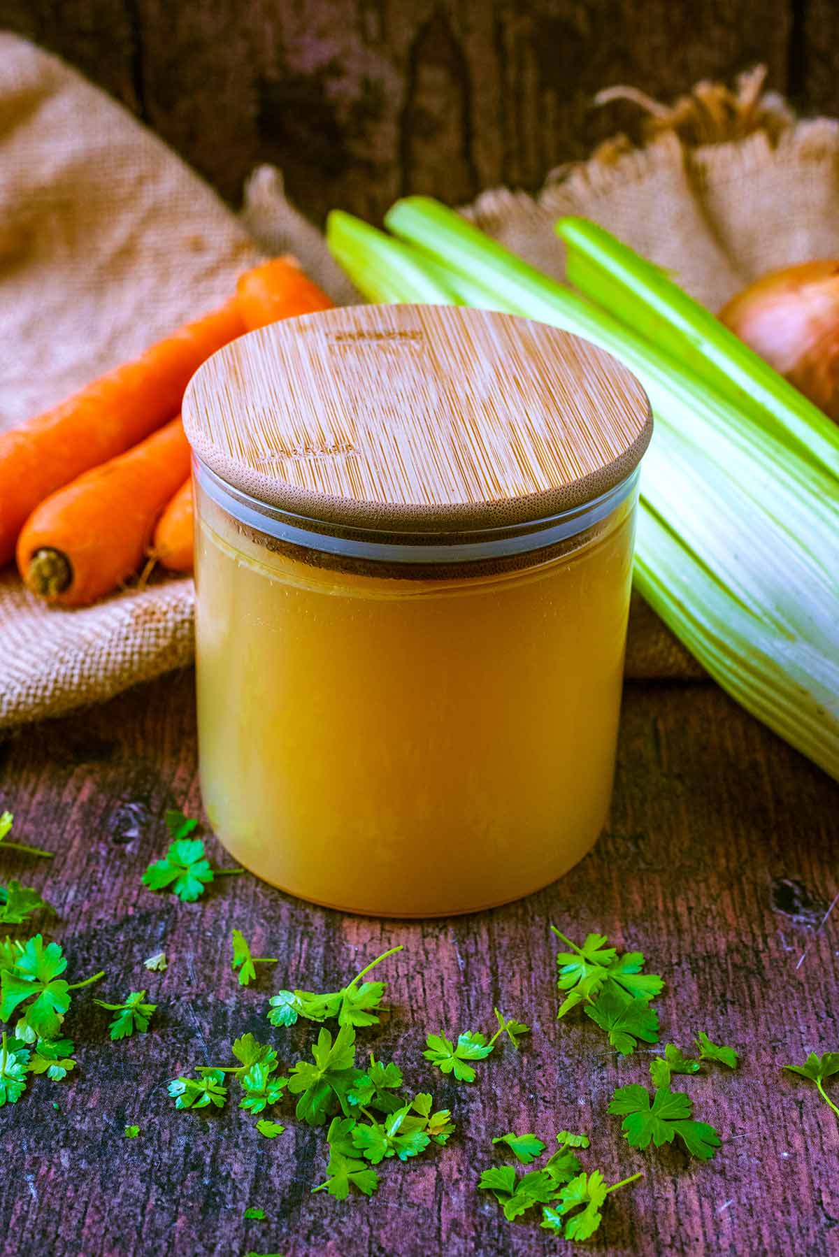 Chicken stock in a jar in front of some carrots, celery and an onion.
