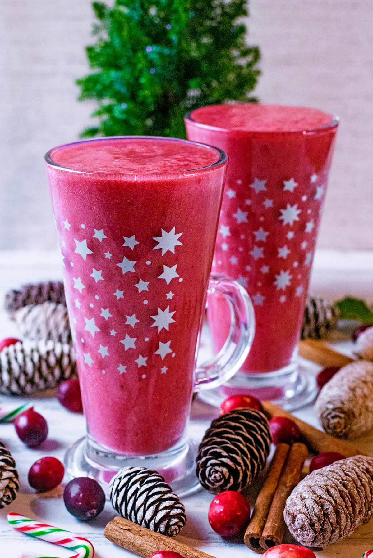 Two glasses containing cranberry smoothie surrounded by pinecones and cinnamon sticks.