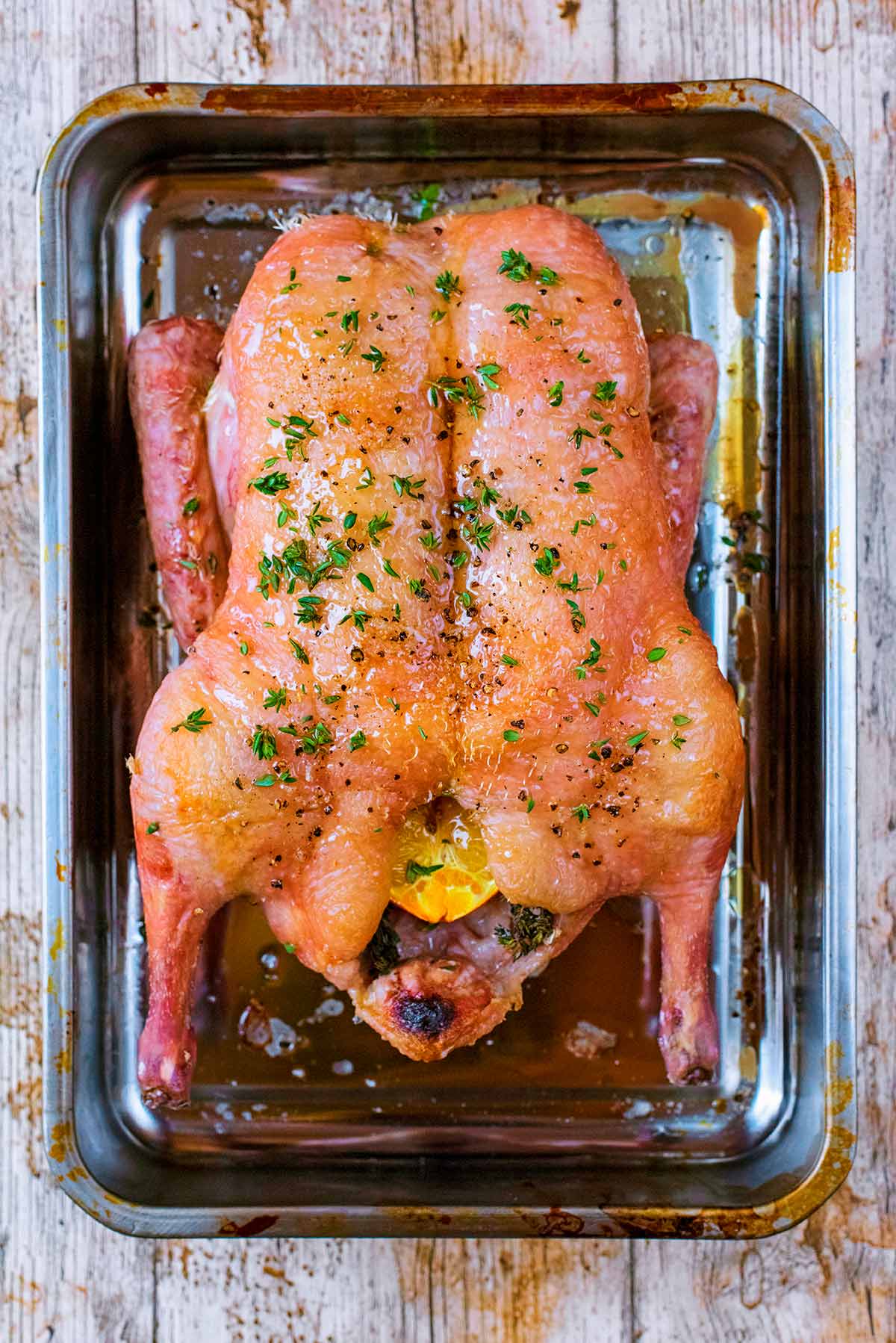 A whole partially cooked duck in a roasting pan with honey and thyme leaves on top.