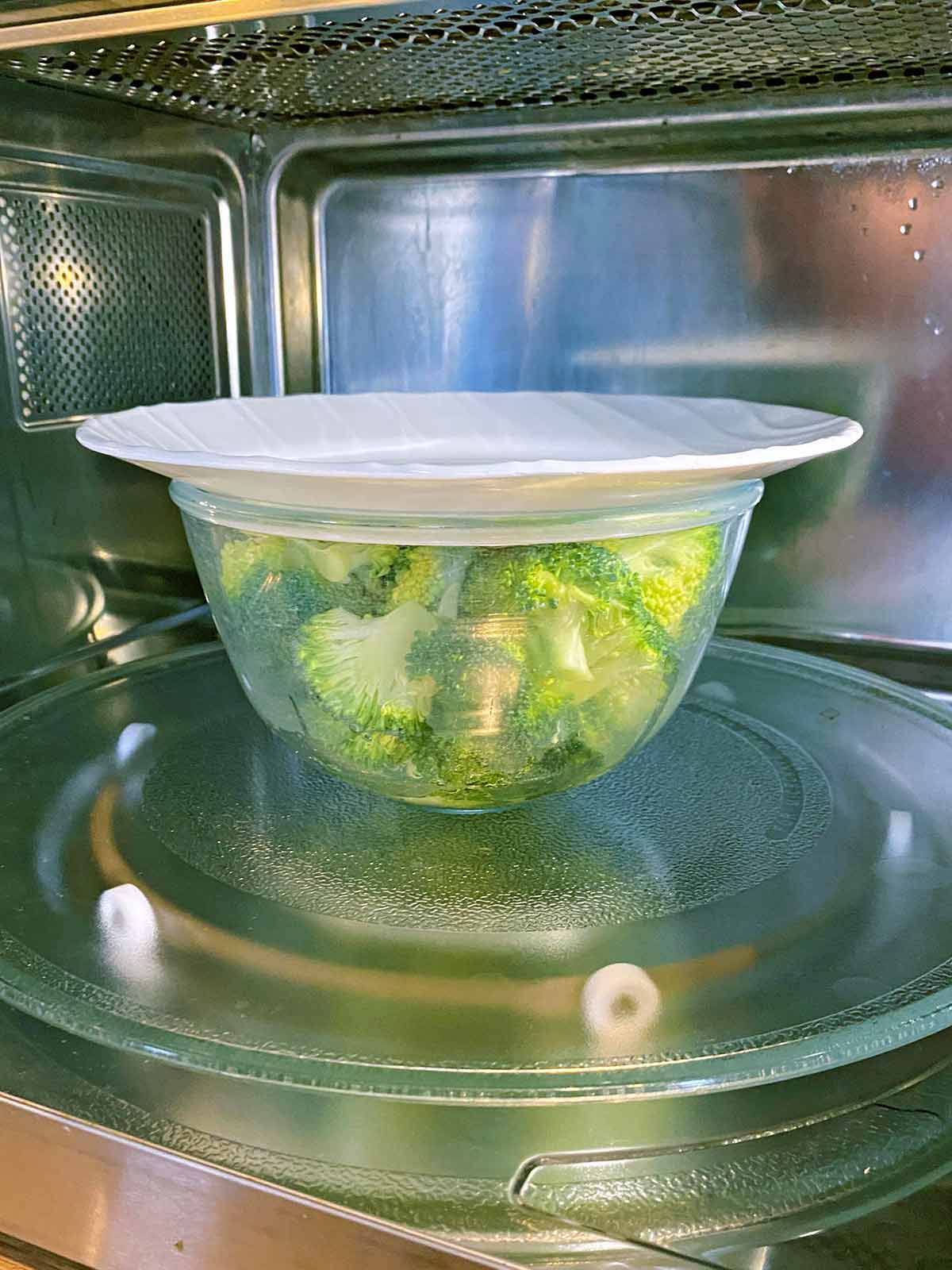 A bowl of broccoli covered with a plate in a microwave.