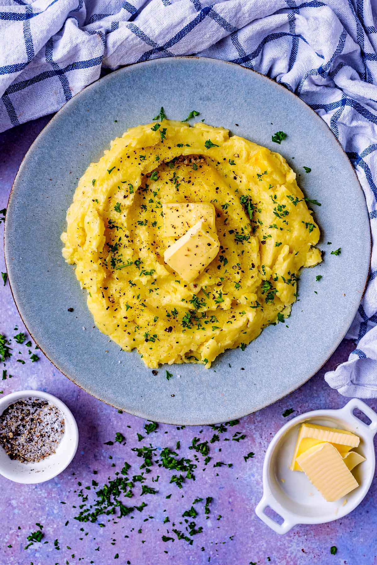 A plate of mashed potato next to a towel, some butter and salt and pepper.