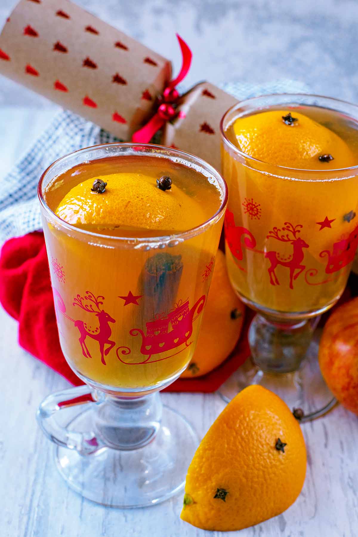 Two glasses of Mulled Apple Juice with orange wedges and a Christmas cracker.