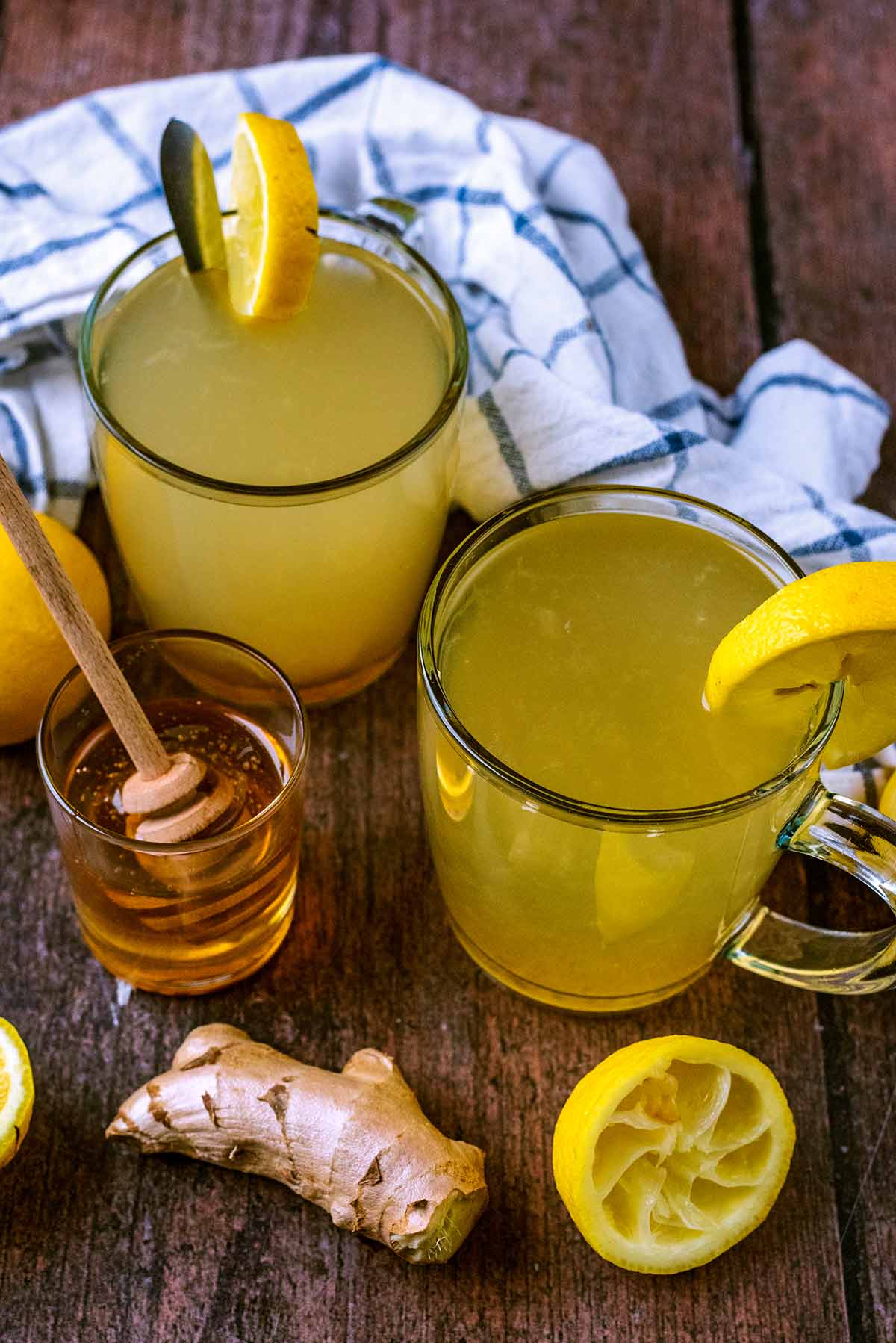 Two glasses of hot lemon drink next to a pot of honey, some ginger and a lemon half.