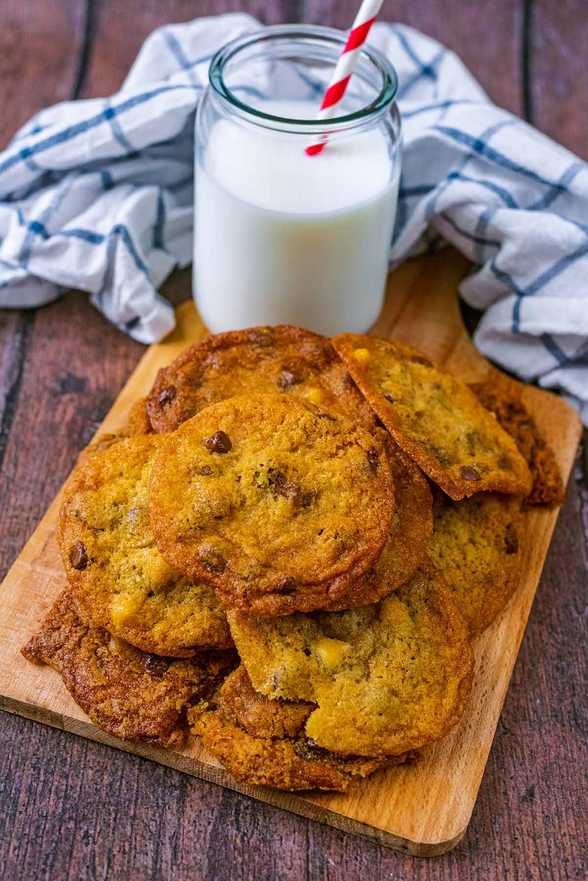 A pile of chocolate chip cookies on a wooden board with a glass of milk.