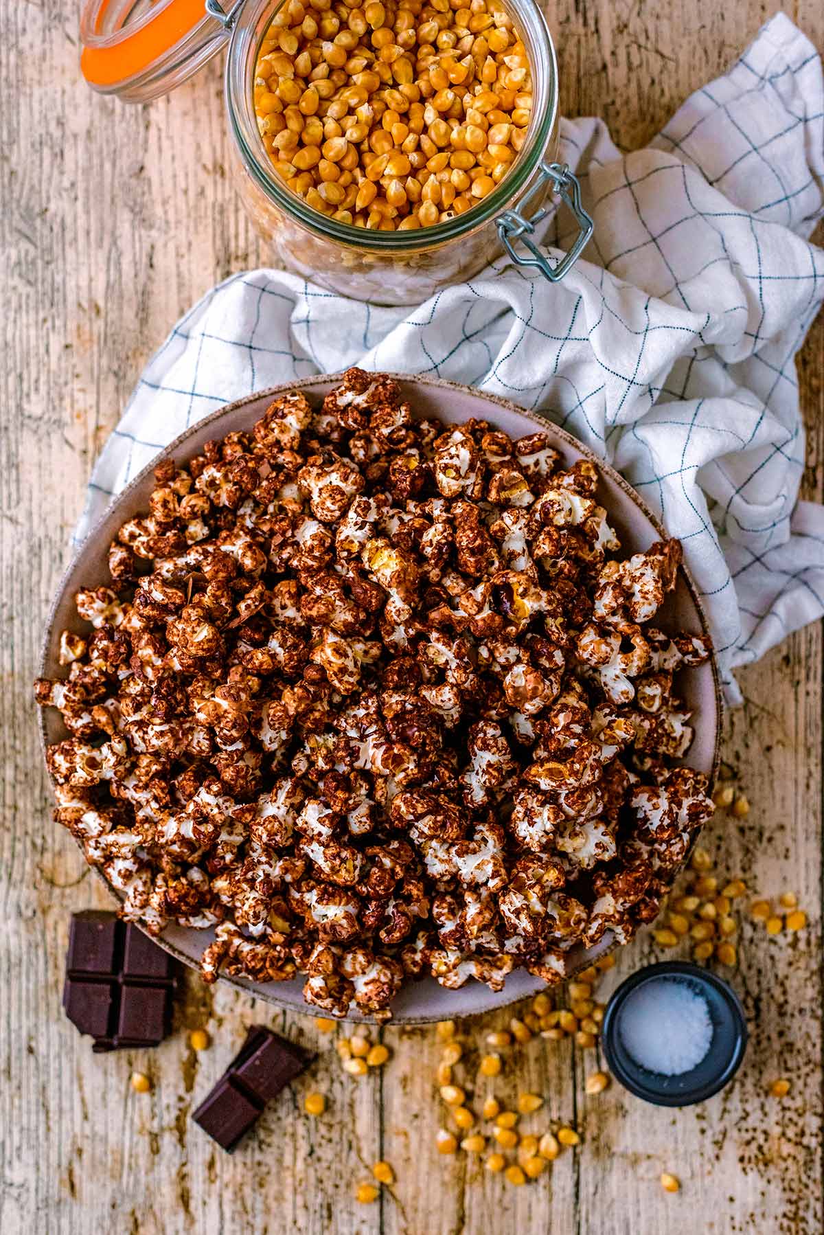 A bowl of chocolate covered popcorn next to some kernels, salt and chocolate.