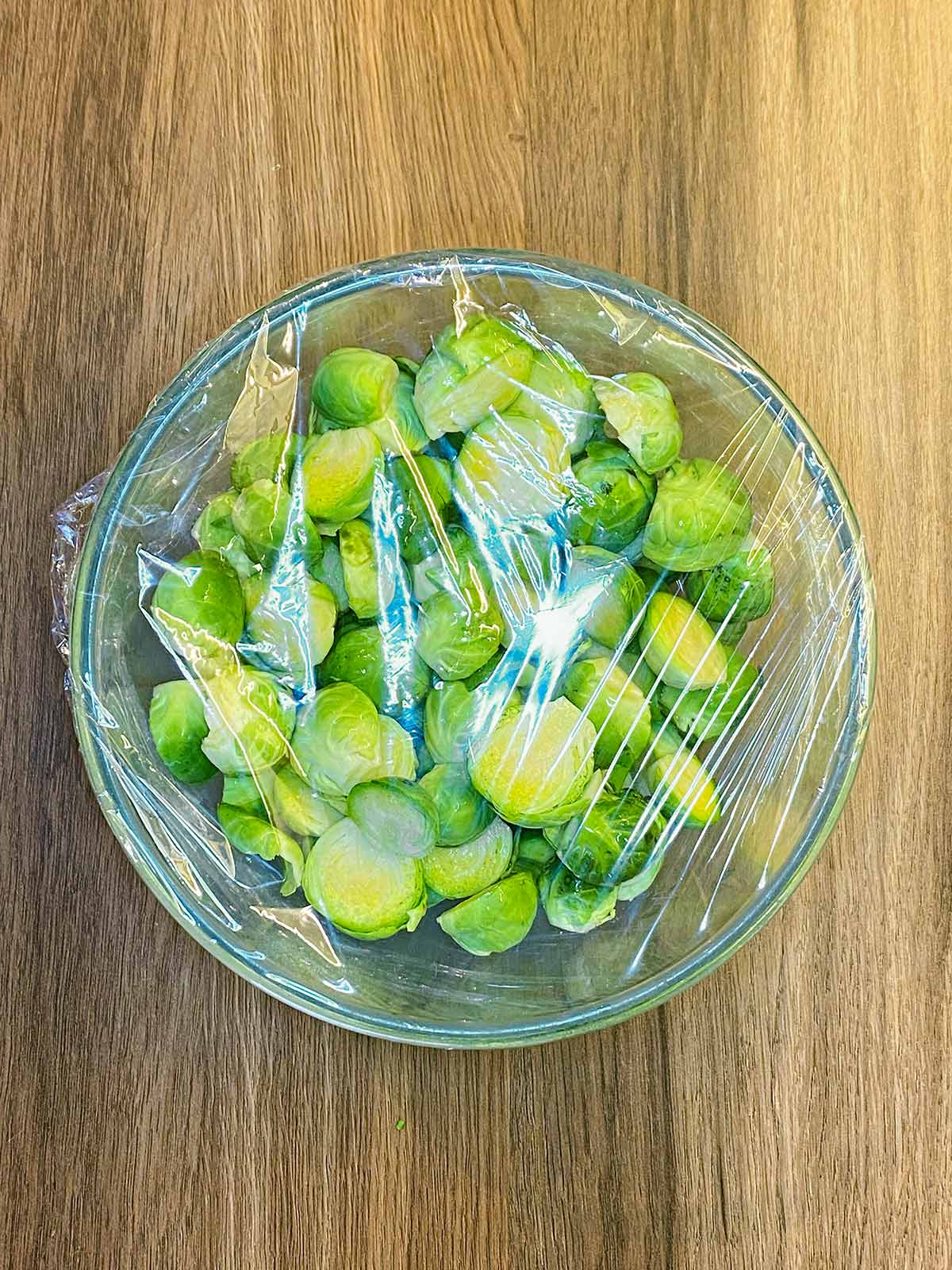Halved sprouts in a glass bowl covered with plastic wrap.