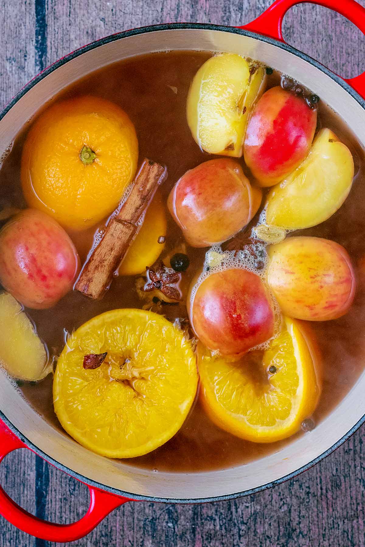 Apples, oranges, cinnamon sticks and spices in a liquid in a pan.