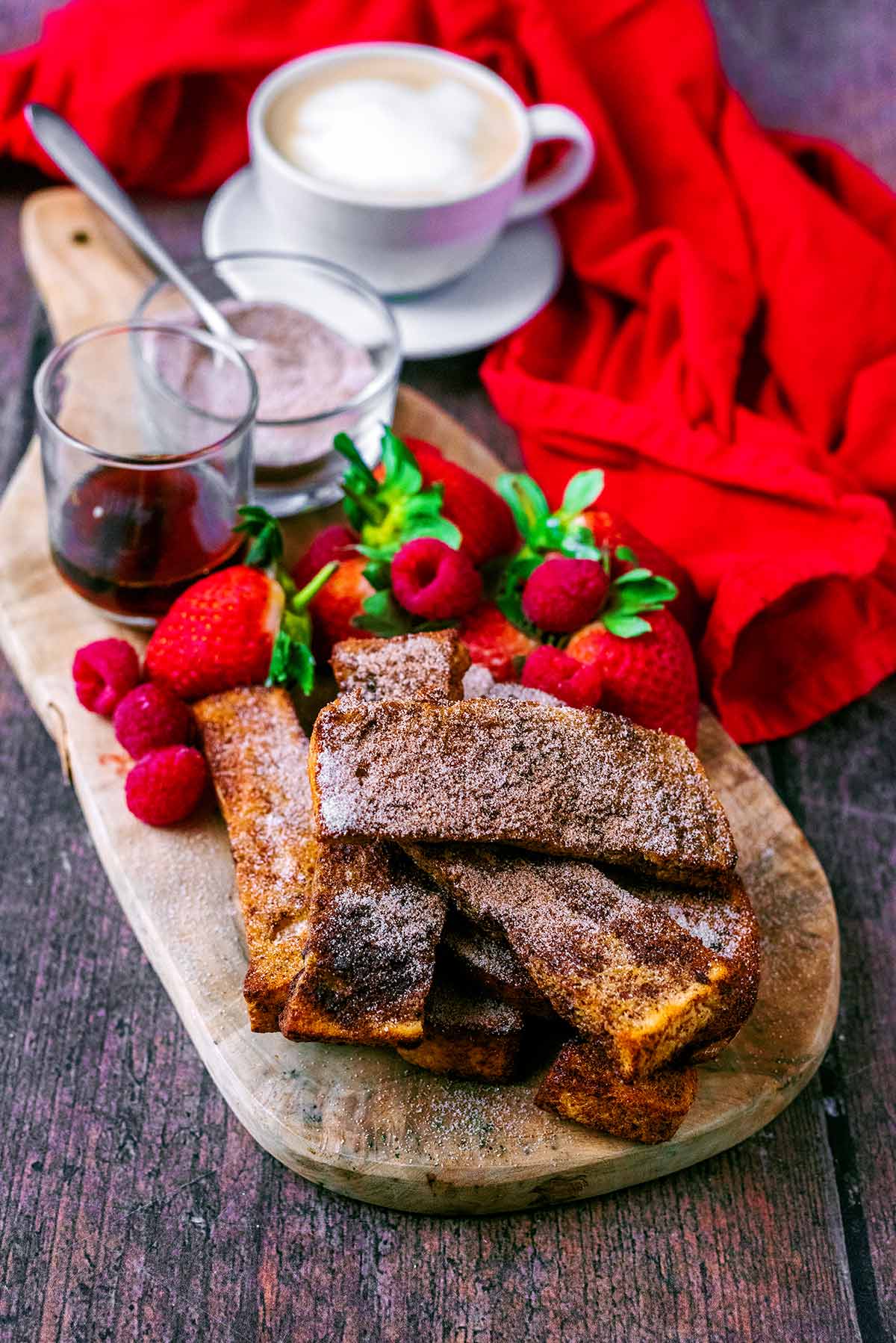 French toast on a board with berries, cinnamon sugar and some maple syrup.