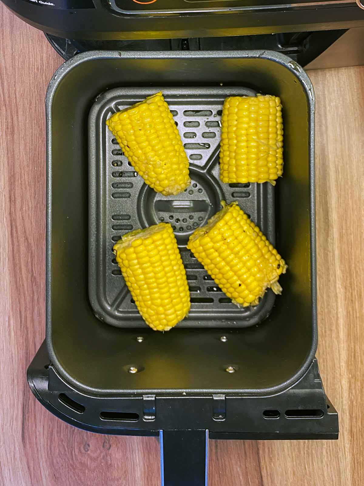 Four uncooked corn cobs in an air fryer basket.
