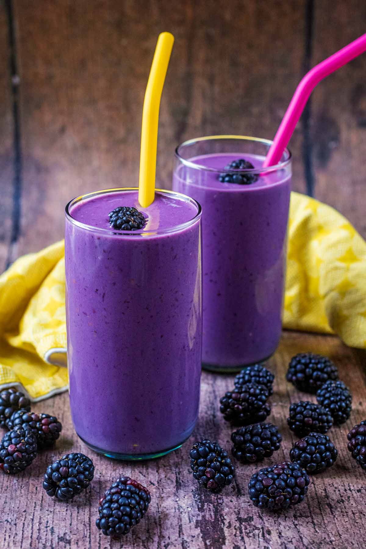 Two purple coloured smoothies in tall glasses with blackberries scattered around.