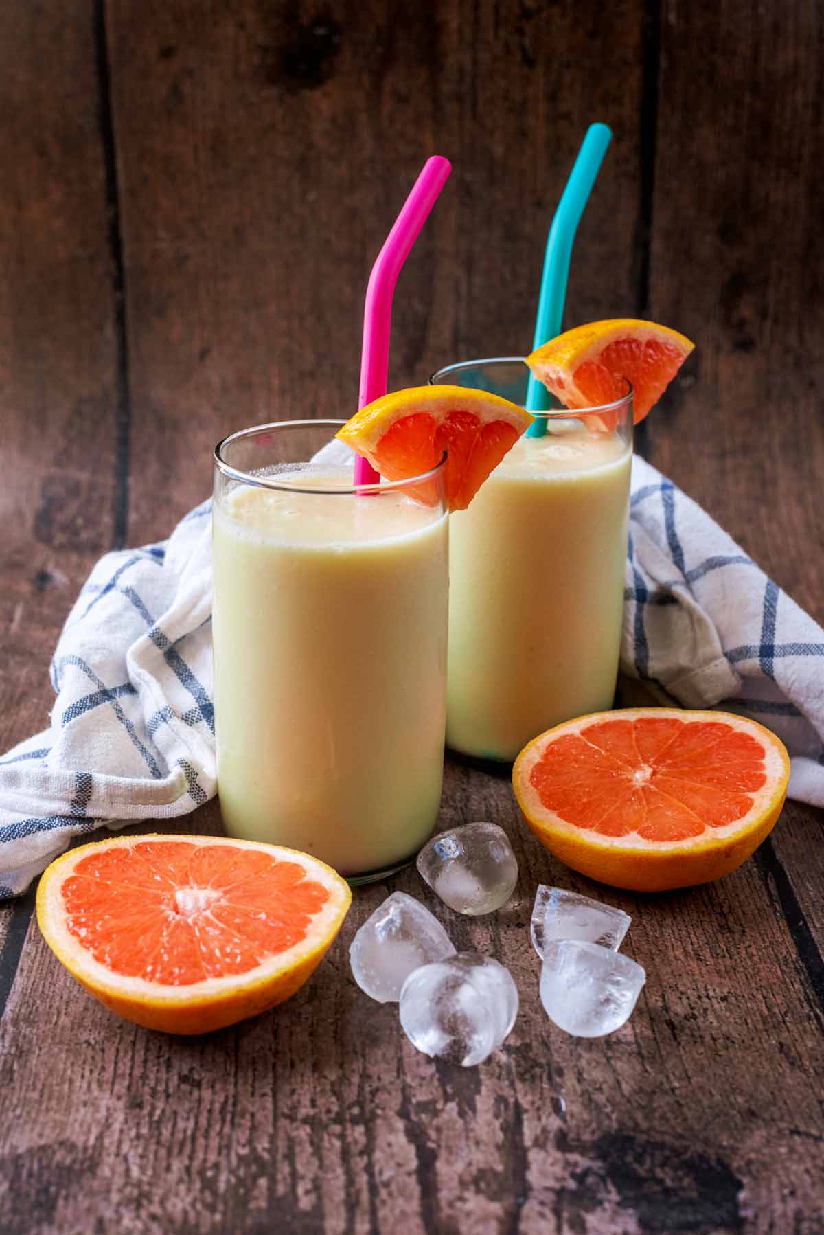 Two glasses of smoothie next to slices of grapefruit and some ice cubes.