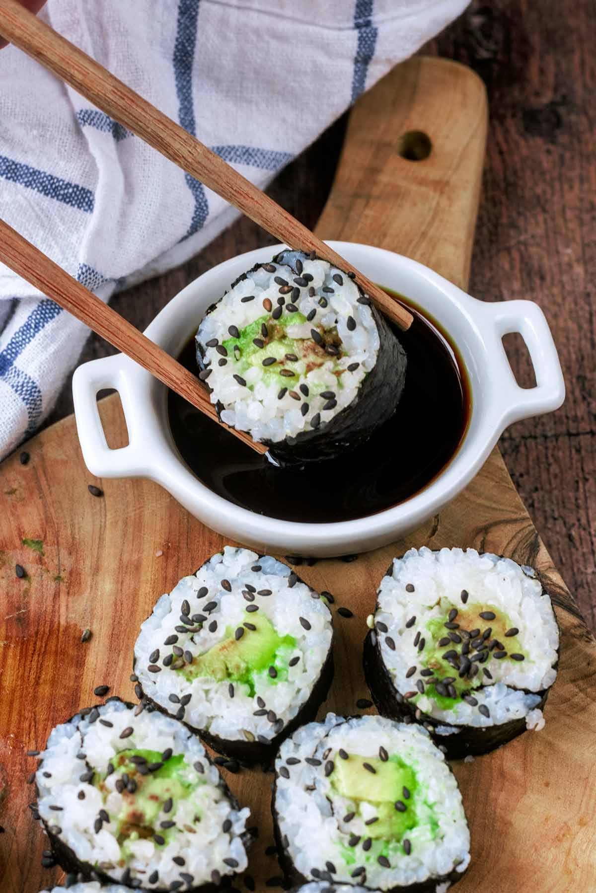 A pair of chopsticks dipping some avocado sushi into a small dish of soy sauce.