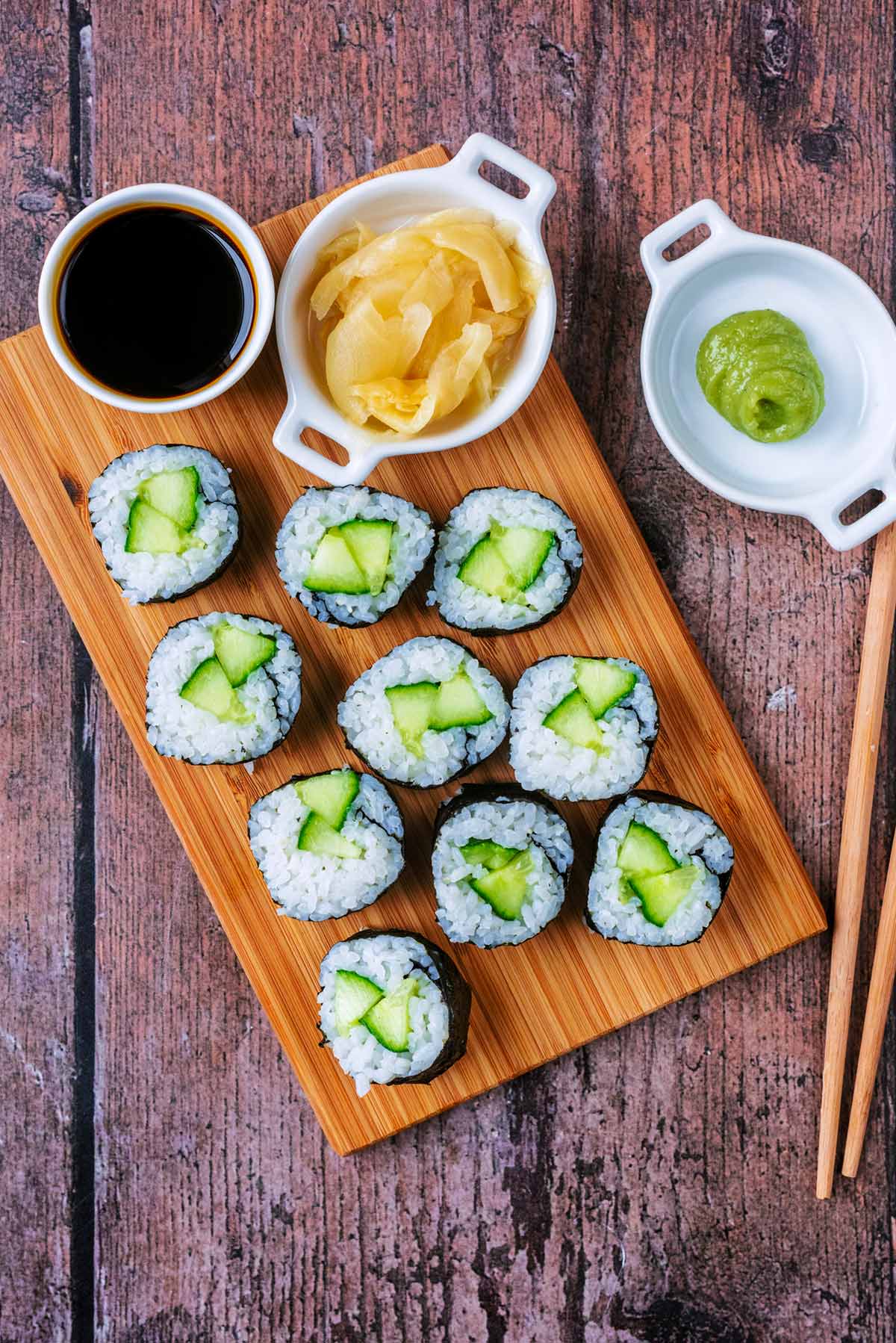 A wooden serving board with cucumber sushi, soy sauce, ginger and wasabi.